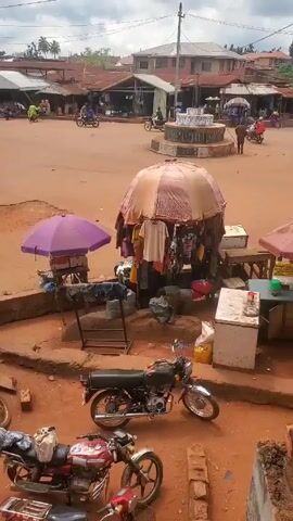 The video shows a wide, unpaved town square or market area with a light-reddish, sandy surface.  There are a few buildings with metal roofs and several makeshift stalls or kiosks scattered around.  One stall in the foreground is shaded by a large, mottled umbrella and displays clothing for sale. A purple umbrella shades a smaller stall nearby. Two motorcycles are parked in the foreground.  In the center of the square is a multi-tiered, circular structure that appears to be a fountain. A few people can be seen walking or standing near the fountain. Several motorcycles are seen driving across the square. Overhead, power lines crisscross the scene against a cloudy sky.