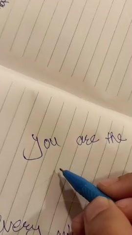 A close-up shot of a person's hand holding a blue pen and writing on a lined notebook. The text being written reads "You are the best OK". The notebook is white with thin blue lines. The pen is blue and has a silver tip. The hand is holding the pen with its index finger and thumb. The lighting is bright and even.