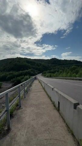 The video begins with a shot of a road and a concrete barrier. It then transitions to a woman walking along a concrete path next to a dam. She is smiling and holding the camera in a selfie-like manner. She is wearing a black t-shirt, a plaid skirt, and a baseball cap. The sky is blue with some clouds. Trees and hills are visible in the background. The dam is a large concrete structure. The woman waves and smiles at the camera before the video ends.