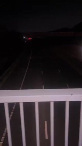 The video shows an elevated view from a bridge overlooking a multi-lane highway at night. White railings of the bridge are visible in the foreground. A large red truck with numerous red taillights and white headlights drives away from the camera in the rightmost lane of the highway. Other vehicles with headlights are visible in the distance traveling in the opposite direction. The surrounding environment is dark, with distant hills or structures outlined against the night sky.