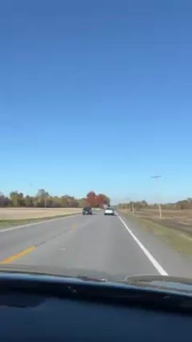 The video shows a view from inside a car driving on a highway. The sky is clear and blue, and there are other cars on the road ahead.  To the right of the road is a field and a row of telephone poles. The field appears to have recently been harvested. The camera pans slightly upward, showing the telephone wires against the clear sky.