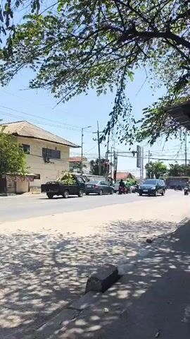 The video shows a street scene with several vehicles and trees. In the foreground, there is a sidewalk with a shadow cast by overhead trees. The street itself is dusty and appears to be an intersection with traffic lights visible in the background. On the street, there are multiple vehicles moving, including a black pickup truck loaded with green vegetation, a dark sedan, a black minivan, and several motorcycles. A few people are visible riding the motorcycles. In the background, there is a building with a light-colored exterior and a red-tiled roof, as well as utility poles and wires. The sky is clear and blue, suggesting a sunny day. The overall impression is a typical urban or suburban street with ongoing traffic.