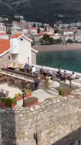 The video shows a high-angle view of a coastal town, possibly in Europe, with stone buildings and orange tile roofs. Several outdoor dining areas are visible, featuring tables and chairs occupied by people. The camera pans, revealing a rocky coastline and turquoise water. Mountains are visible in the background. People are sitting and standing around the seating areas. The scene is bright and appears to be daytime.