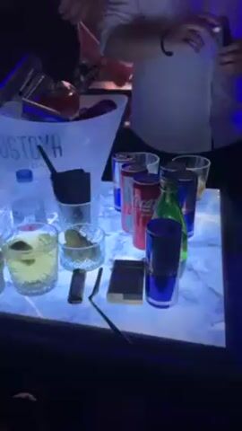 The video shows a well-lit table at what appears to be a nightclub or bar. The table has a light-up surface with a marble pattern. Various items are on the table, including an ice bucket with an “Ostoya Vodka” label containing several bottles, cans of soda and Red Bull, glass bottles (one green), glasses with liquid in them (some with slices of fruit), stacks of coasters, a lighter, and a pack of cigarettes. Several people are gathered around the table, casually dressed and interacting. Some of them raise their glasses in a toast. The background is dark with hints of other people and lighting, indicating a social gathering in a nightlife environment. A man with a close haircut is visible in the foreground. Upbeat music can be heard in the background.