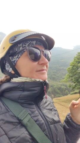 A woman wearing a yellow helmet and sunglasses rides on the back of a scooter being driven on a road. A man in a purple helmet is driving the scooter. There is a mountainous background behind them. The houses are mostly low-rise, and the flag of Vietnam is visible. The woman wears a black jacket with an olive-green strap across her chest.