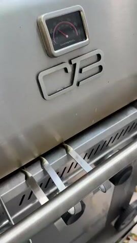 The video shows a close-up view of a grill with food cooking. On the right side of the grill, there are two large steaks, one of which appears to be a ribeye cut. They are searing and have grill marks. On the left side of the grill, there are five skewers of meat, possibly chicken or pork, that are also cooking and appear to be seasoned with a reddish spice. Flames are visible under the meat, indicating active grilling. The grill itself is a stainless steel gas grill with control knobs visible at the bottom. The overall lighting suggests an outdoor setting, likely during the daytime.