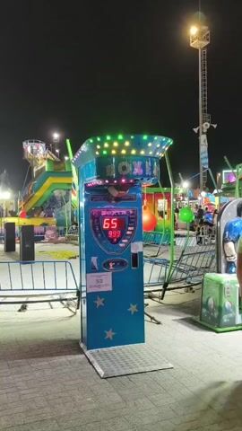 At night, in an amusement park, a boxing game machine stands in the foreground. The machine is blue with colorful lights on top and a digital display showing "65" and "999". A man in a white t-shirt and black shorts stands to the right of the machine, facing it. Behind the machine, other amusement park rides and attractions are visible, including a Ferris wheel and a roller coaster. The ground is paved with interlocking stones.