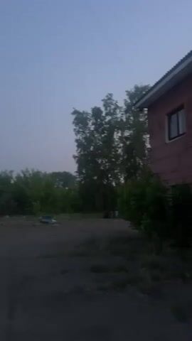 A handheld, low-light video pans to the right. In the foreground, there is a cracked asphalt surface. To the right, a brick building with a window is visible. Beyond the asphalt, there are trees and bushes. In the distance, a metal tower with two speakers is visible against a hazy, pale blue sky. A streetlamp is also visible. There are no people or animals visible in the video.