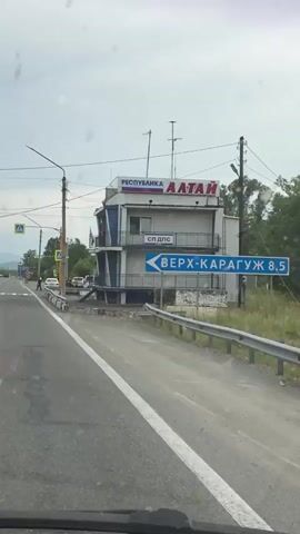 The video begins with a view from a vehicle of a multi-story building on the right side of the road. The building has signs in Russian, including "РЕСПУБЛИКА АЛТАЙ" and "СП ДПС". A blue directional sign reads "ВЕРХ-КАРАГУЖ". A pedestrian crossing with yellow and white stripes is visible on the road. There are traffic lights and streetlights. Several cars are parked near the building and on the side of the road. Some people are visible near the pedestrian crossing and on the sidewalk. The road appears to be a highway or a major road with multiple lanes. The environment appears to be a rural or semi-rural area with trees and hills in the background. The sky is overcast. The vehicle continues to move forward, passing the pedestrian crossing.