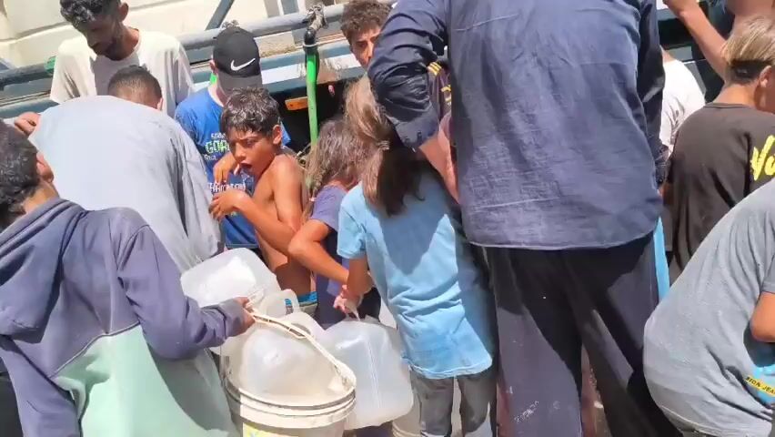 The video shows a group of children and adults gathered around a water distribution point. Several children, some shirtless, are holding empty plastic water jugs. One boy in a black and yellow soccer jersey is speaking animatedly. Another boy wearing a blue t-shirt and a black baseball cap appears to be assisting with the water distribution. The environment looks like an outdoor urban setting, possibly during the daytime, with a truck or vehicle in the background that appears to be the source of the water. The overall scene depicts people waiting to collect water, suggesting a situation where water access may be limited.