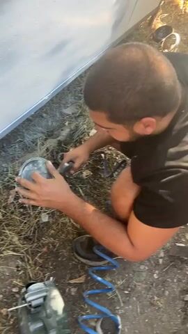 A person is crouched down outdoors, holding a spray gun. They are detaching a cup from the spray gun. The cup appears to contain a light-colored liquid. The ground is covered in dry grass and dirt. A blue coiled hose is visible on the ground. In the lower left corner, a green jerry can is present.