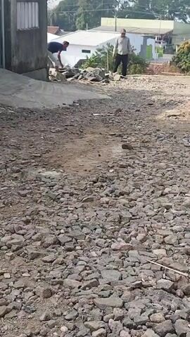 Two men are working on a dirt and gravel road. One man in a blue shirt and light-colored pants is bending over, appearing to be moving or arranging rocks. The other man, wearing a light-colored long-sleeved shirt and dark pants, is also bending and appears to be working with rocks. The environment is outdoors, with a building on the left, and other structures and trees visible in the background. The road is uneven and covered with loose stones and dirt. The lighting suggests it is daytime.