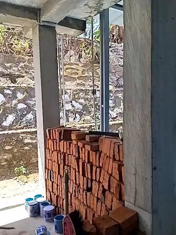 The video shows a construction site. In the foreground, a stack of red bricks is visible next to a concrete pillar. Further back, a terraced stone wall is seen with vegetation growing on it. Two men are sitting on chairs in the background, facing away from the camera. The ground is covered in blue concrete, and a roof with metal sheeting is overhead.
