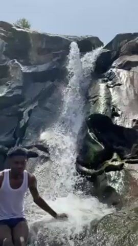 The video shows a young man sitting on wet rocks in front of a small waterfall. The man has dark skin, short dark hair, and is wearing a white tank top and dark shorts. He looks into the camera and says something, but the audio is unclear. He uses his hands for emphasis while he speaks. The waterfall cascades down a dark gray rock face, creating a pool of turbulent white water at its base. The surrounding rocks appear to be wet and moss-covered. The sky is visible above the waterfall.