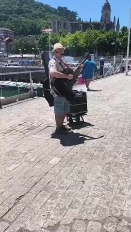 A man with a hat and sunglasses is playing an acoustic guitar on a cobblestone path next to a body of water with boats. He is wearing a short-sleeved shirt, shorts, and sneakers. His guitar is in a case, and he has a small amplifier on a cart behind him. In the background, there is a building with a tall spire, trees, and a hill. Another person is walking away in the background. The sky is blue and the sun is shining.