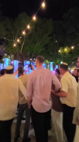 The video shows a group of men dancing outdoors at night. They are wearing yarmulkes and white shirts, and appear to be holding hands in a circle. There is a stage with a light display in the background, and string lights are hanging overhead. The overall atmosphere seems festive.