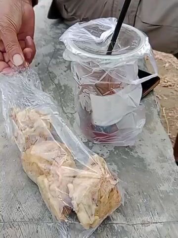 The video shows a close-up of a person's hands reaching for a plastic bag containing several pieces of fried food, possibly chicken, which are arranged in a rectangular shape. The food appears golden brown and slightly greasy. Next to the bag of food is a clear plastic cup with a dark liquid and a black straw, also contained within a plastic bag. The cup is placed on a rough, gray surface, possibly a table or a bench, which is outdoors. The background is blurred, but suggests a sandy or dry ground. The person's hands are visible in the frame, with one hand holding the plastic bag of food and the other hand, with visible wrinkles, appears to be reaching out. The overall scene suggests a casual outdoor setting where food and drinks are being prepared or consumed.
