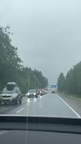 This is a first-person perspective video captured from inside a vehicle on a highway in Razbegayevo, Russia. The weather is overcast and appears to be raining, as evidenced by the wet road surface reflecting the headlights of the cars and the motion of the windshield wipers. The scene depicts a significant traffic jam, with vehicles lined up as far as the eye can see on the highway. The surrounding environment is characterized by dense green trees lining both sides of the road, suggesting a rural or semi-rural setting.

The primary objects in the scene are numerous cars and a few vans and trucks, all stationary or moving very slowly. The vehicles are varied in make and model, and several have their headlights on due to the poor visibility. The video focuses on the progression of the traffic jam, with the view shifting slightly as the camera vehicle inches forward. The overall mood conveyed is one of frustration or patience, common in such traffic situations.

No people are clearly visible outside the vehicles, and the focus remains on the road and the traffic. There are no explicit time-of-day indicators, but the overcast sky and headlights suggest it is daytime, possibly late afternoon or early evening. Some license plates are visible, but the text is not clear enough to read specific details, except for what appear to be Russian characters and numbers. A blue highway sign is visible in the distance, but the text on it is illegible.