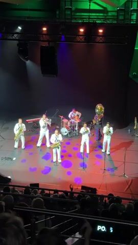 The video shows a band performing on a stage in front of an audience. The band consists of eight musicians playing various instruments. Four of the musicians are playing saxophones, one is playing a keyboard, one is playing the drums, and one is playing a tuba. The musicians are all wearing white uniforms. The stage is lit with purple and red spotlights. The audience is seated in a theater, with the seats visible in the foreground. The video is recorded from a distance, providing an overall view of the performance.
