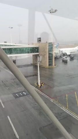 The video shows an airport tarmac during a rain shower. A jet bridge extends from the terminal to an airplane. Several vehicles, including a green van and a baggage tug, are on the tarmac. The ground is wet, and raindrops are visible on the window through which the footage is being recorded. The sky is overcast and hazy.