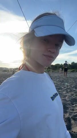 The video is a selfie taken by a young person with freckles, wearing a white long-sleeved shirt and a blue visor. They are on a sandy beach with a volleyball net in the background. The sun is shining brightly, creating lens flare. In the first part of the video, the person is looking at the camera and smiling. In the second part, they close their eyes and tilt their head. In the third part, they lean into the camera and pucker their lips. In the background, there are other people on the beach.