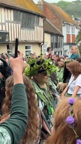 A crowd of people gathers on a narrow street in a European town. It is a pedestrian zone. Many are dressed in costumes with a folkloric, pagan theme. Many of the costumes are green and floral and include foliage on hats and clothing. One person wears a black animal mask with large horns. A child sits on someone’s shoulders. A wooden staff decorated with feathers is seen. The background includes houses and street signs.
