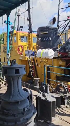 The video shows the stern of a yellow tugboat, with the number CPA-3069 visible on a black structure. A white life raft is attached to the side of the tugboat. Various pieces of equipment, including winches and ropes, are on the deck. In the background, there are buildings and a body of water. The sky is blue with some clouds. The camera pans slightly to the right, revealing more of the tugboat and the surrounding environment.