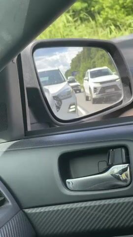 The video captures a close-up view from inside a car, looking at the side-view mirror. The mirror reflects a highway scene with several cars. The primary focus is on the white SUV directly behind the car the video is being filmed from, visible in the mirror's center. To its right, another white SUV is further down the road. A dark-colored vehicle is also visible in the distance on the highway.

The setting appears to be a sunny day, with bright light and clear skies reflected in the mirror. Lush green trees line the side of the highway, indicating a rural or suburban location. The car's interior is visible in the foreground, featuring a textured door panel and a chrome door handle.

The activity is the cars driving on a highway. There's no overt indication of any specific event, but the proximity of the vehicles suggests normal traffic conditions. The overall impression is one of a typical road trip or commute. No text is visible in the media. The location context of Zanevka, Russia, places the scene geographically.