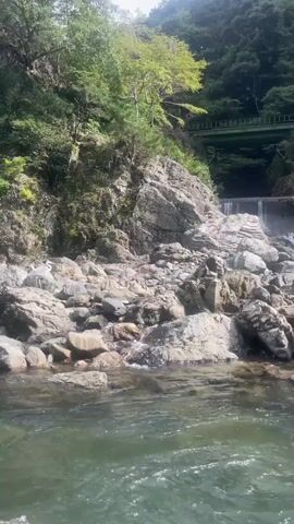 A raft navigates a rocky river under a bridge and surrounded by trees. Two people are on the raft, one in front and one at the back. The person in the back is wearing a cap and looking forward, while the person in front is wearing a hat and a white t-shirt, holding an oar and appearing to speak.