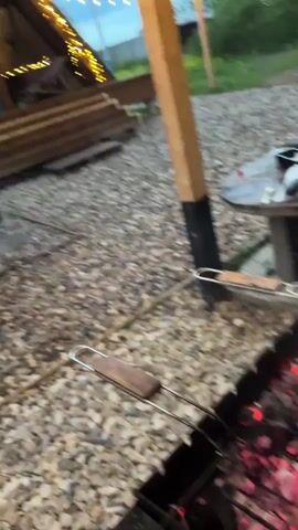 The video shows an A-frame house with lights along the top of its frame. The house is behind a charcoal grill full of burning embers and meat grilling over them. The ground is covered in small gravel.