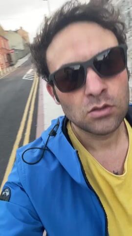 A man wearing sunglasses and a blue jacket over a yellow shirt is walking down a street. The camera is held at a low angle, and the man's face is the primary focus. The background shows a stone wall on the right and a street with buildings on the left. The video appears to be a selfie video recorded while walking.