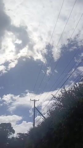 The video shows a view of the sky from below, framed by the branches of a tree and crossed by several power lines. The sky is a mix of blue and white, with fluffy clouds scattered throughout. The camera moves slightly, shifting the perspective of the clouds and revealing more of the blue sky in certain areas.