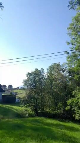 The video shows a landscape view from a moving vehicle. The sky is clear blue with some wispy clouds. In the foreground, there are green grassy fields and trees. In the background, there are rolling hills and mountains. Some houses and buildings are visible on the hillsides. Power lines are visible against the sky. The video appears to be taken during the daytime, likely in the summer or spring, given the lush green vegetation.