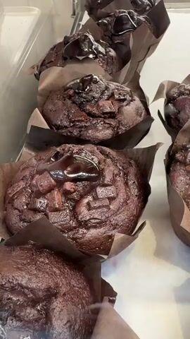 This media file shows a close-up, overhead view of a display of freshly baked chocolate muffins. The muffins are dark brown, rich in color, and appear to be topped with chocolate chips and a drizzle of glossy chocolate sauce. They are presented in brown paper wrappers, arranged in rows on a white surface, possibly a display case in a bakery or cafe. The lighting suggests an indoor setting, possibly during daylight hours.

In the background, slightly out of focus, are what appear to be glazed donuts with a topping of nuts or other crunchy pieces. The scene is clean and appealing, focusing on the tempting appearance of the baked goods. There is no visible text in the media file. The overall impression is one of delicious, indulgent treats ready for sale. Given the context of Adeje, Spain, this could be a bakery or patisserie serving local residents and tourists.