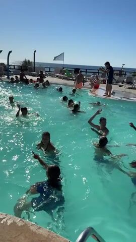 A group of children and adults are in a swimming pool on a sunny day. Some are swimming, while others are standing around the edge of the pool. Two people are standing on the edge of the pool, one holding a phone and appearing to record. A flag is visible on a pole in the background, along with lounge chairs and the ocean. The water is clear and blue, and the sky is clear and blue. Some of the people in the water are waving and interacting with each other.