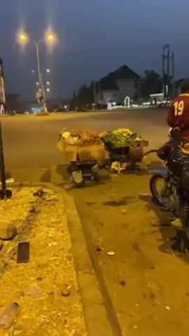 The video shows a street scene, probably at dusk or dawn, with street vendors selling food or produce. There are two wheeled carts, one with what appears to be fried food, the other with fresh produce like carrots and lettuce. A man sits on a low stool, possibly preparing food. Another man in a sports jersey walks past. A motorcycle is parked nearby. There is a sign with phone numbers for a school. The street is mostly empty, with some distant cars visible. The environment looks urban, possibly a busy intersection. There is litter on the ground.