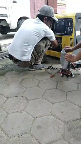 Two men are crouched on a hexagonal paved surface, working on a yellow generator. The man on the left, wearing a white t-shirt, khaki pants, a grey baseball cap, and glasses, is examining the interior of the generator, which has a panel open. Tools are scattered on the ground between the two men, including screwdrivers, pliers, and wires. The man on the right, wearing a dark t-shirt and a baseball cap with an orange brim, is holding a gray cloth and seems to be assisting. A white truck is visible to the left of the generator, and a brick wall is behind them. The overall scene suggests they are performing maintenance or repairs on the generator.