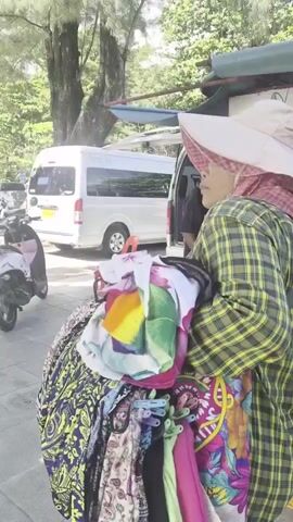 The video shows a person from the shoulders down, wearing a wide-brimmed hat and a plaid shirt, carrying a large bundle of colorful fabrics. The person is walking on a paved walkway next to a sandy area with trees. In the background, there is a white van, a parked motorcycle, and a food cart with people around it. The scene appears to be outdoors, possibly in a tropical or beachside location. The lighting suggests it is daytime.