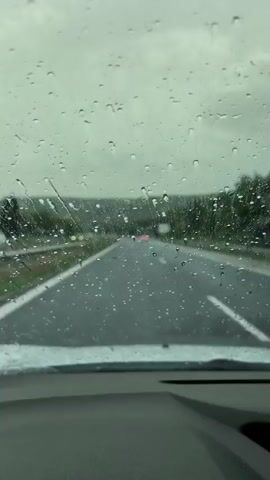 The video shows a view from inside a car on a highway during rainfall. Raindrops are visible on the windshield, distorting the view of the road ahead. The road is wet, and there are other vehicles in the distance. The sky is overcast. The car appears to be moving forward.