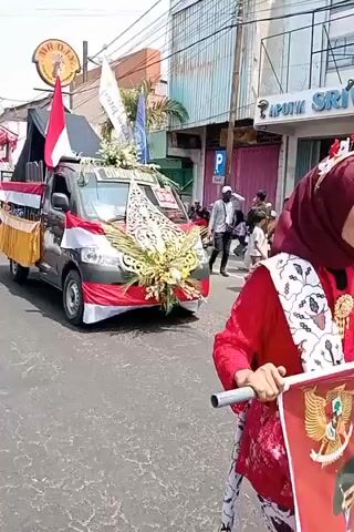 A dark gray pickup truck decorated with Indonesian flags and floral arrangements is proceeding down a paved street. The truck has a large black tarp covering the rear bed. The side of the truck bed is adorned with gold fabric. The front of the truck is decorated with red and white fabric, resembling the Indonesian flag, and elaborate floral and leafy arrangements. A large gold, ornate, carved wooden piece is attached to the front of the truck. The truck appears to be part of a parade or procession. Several people are visible on the sidewalk to the right of the truck, some are seated, and some are standing. One person, dressed in a red hijab, is walking behind the truck. Another person in a dark shirt and white head covering is standing near the sidewalk. Buildings line the street on the right, with signage including "APOTIK SRIPATI." The sky is bright, indicating daytime. The overall scene suggests a festive or celebratory event.