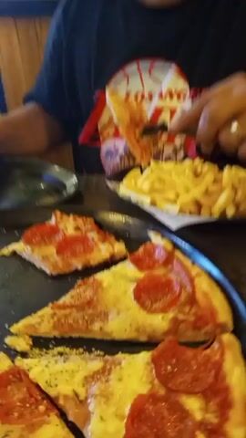 A close-up shot shows a black table with a black pizza tray filled with partially eaten pepperoni pizza slices and a pile of french fries on a small plate. A person wearing a black t-shirt with a red and white design uses a black plastic fork to pick up a piece of pizza. Another person's hand reaches toward the pizza tray. The scene appears to be inside a restaurant or dining area.