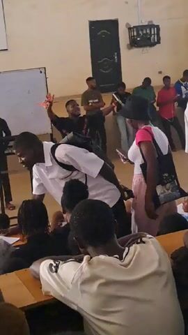 The video shows a classroom with students sitting at desks. A man wearing a white shirt and black backpack is leaning over desks, interacting with students. Some other people are standing in the back of the room. The setting appears to be a university or college lecture hall, indicated by the rows of desks, a whiteboard, and the age of the people present. The general atmosphere seems relaxed, with students chatting and moving around.