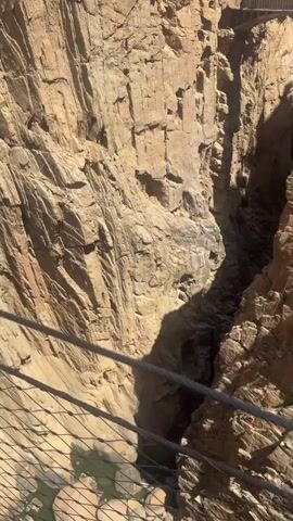 A video shows a deep gorge with steep, rocky walls. A narrow pedestrian bridge spans the gorge, with several people visible on it. The bridge is suspended high above the gorge floor. The sun is shining, casting shadows on the rock faces.