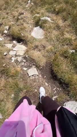The video shows a person walking on a grassy mountain trail. The camera is held by the person, looking down at their feet and then panning up to reveal the mountainous landscape. Other hikers are visible in the distance. The terrain is rocky and covered in dry grass, with a clear blue sky and some clouds. The person is wearing a pink hoodie and dark leggings, and is carrying a backpack. They are also using trekking poles to assist them on the hike.
