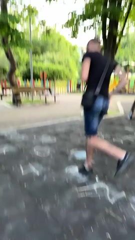 A young man wearing a black t-shirt, denim shorts, and sneakers plays hopscotch on a playground. He has a black shoulder bag slung across his body. The playground has a rubberized surface with chalk drawings of hopscotch numbers. In the background, there are colorful benches, trees, and playground equipment. The man is shown hopping from one square to another, demonstrating agility and coordination. The sun is shining, casting shadows on the ground.