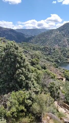 The video showcases a scenic view of a lake surrounded by lush green hills under a partly cloudy sky. The camera pans slowly, revealing the lake's calm, dark waters and the dense vegetation covering the hillsides. A wooden staircase and viewpoint platform is present in the foreground, adding depth to the landscape. An electrical tower stands to the right side of the lake. The overall impression is that of a tranquil, natural setting, offering a peaceful vista.
