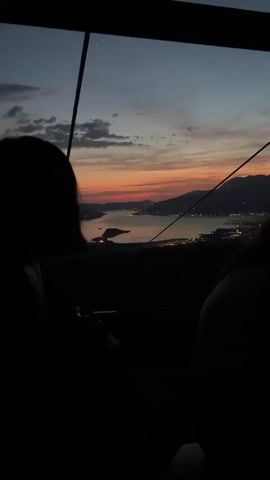 The video captures a scenic view from inside a moving cable car during sunset. Two individuals are visible in silhouette, seated with their backs to the camera, looking out the large window. The sky displays a beautiful gradient of orange, pink, and dark blue as the sun sets behind distant mountains. Below, a body of water reflects the last light of day, bordered by a town whose lights are beginning to twinkle. The cable car's internal structure is dark, framing the vibrant outdoor scene.