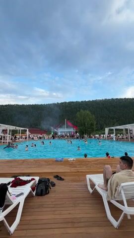 The video opens with a wide shot of a crowded outdoor pool area on a cloudy day. In the foreground, two empty white lounge chairs sit on a wooden deck. A man is visible on the right lounge chair, partially obscured by a towel. A backpack, shoes, and clothing are on the left lounge chair. The pool is filled with people swimming and socializing. In the background, a large tent with red accents and a stage is set up, with smoke emanating from it. A wooded hillside is visible behind the tent. The video pans slightly, showing more of the pool area and the surrounding landscape. A woman in a bikini is seen on the left side of the frame, drinking from a red cup. Several other people are walking around the deck and in the pool. The overall impression is of a lively outdoor event.