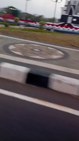 The video shows a blurry, fast-moving shot of an outdoor scene. In the background, a building resembling a mosque with a golden dome is visible. It is flanked by what appear to be fountains or water jets. A decorative border with red, white, and blue colors runs along the front of the area. In the foreground, a road with white markings and black and white barriers is present. The motion blur suggests the video was taken from a moving vehicle. The overall environment appears to be a public space or plaza, possibly during the day or twilight.