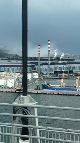 The video shows a wide view of a large industrial port facility on a cloudy day. The scene is viewed from an elevated position. At the center are two tall smokestacks painted with red and white stripes, emitting plumes of white smoke. To the left of the smokestacks is a cluster of white storage tanks and cranes, and on the right a large ship is docked at the pier. In the background, the large industrial buildings of the port stretch along the horizon, with hints of distant hills. The sea is choppy and gray. A metal railing is visible in the foreground, indicating the viewer is on a bridge or elevated platform. There are no people visible in the video.